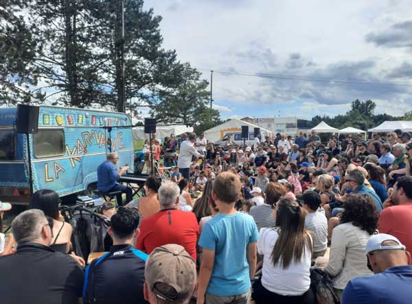 Public nombreux en plein air assistant à une représentation avec une personne jouant devant une caravane bleue 