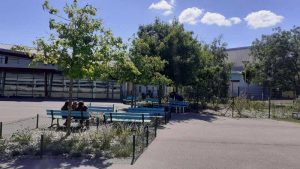 Cour de collège avec des arbres et deux collégiens assis sur un banc avec le bâtiment du collège au second plan